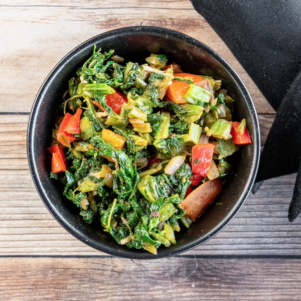 A black bowl filled with African collard greens, red bell peppers, and onions on a wooden table.